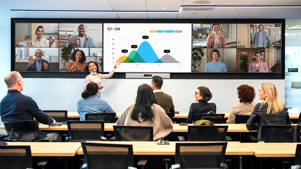 A hybrid meeting room with in-person participants and remote attendees displayed on a large video wall, alongside a shared presentation