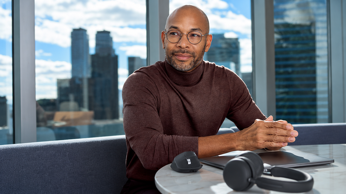 A man wearing glasses and a burgundy sweater sits at an office table with headphones and a laptop beside him