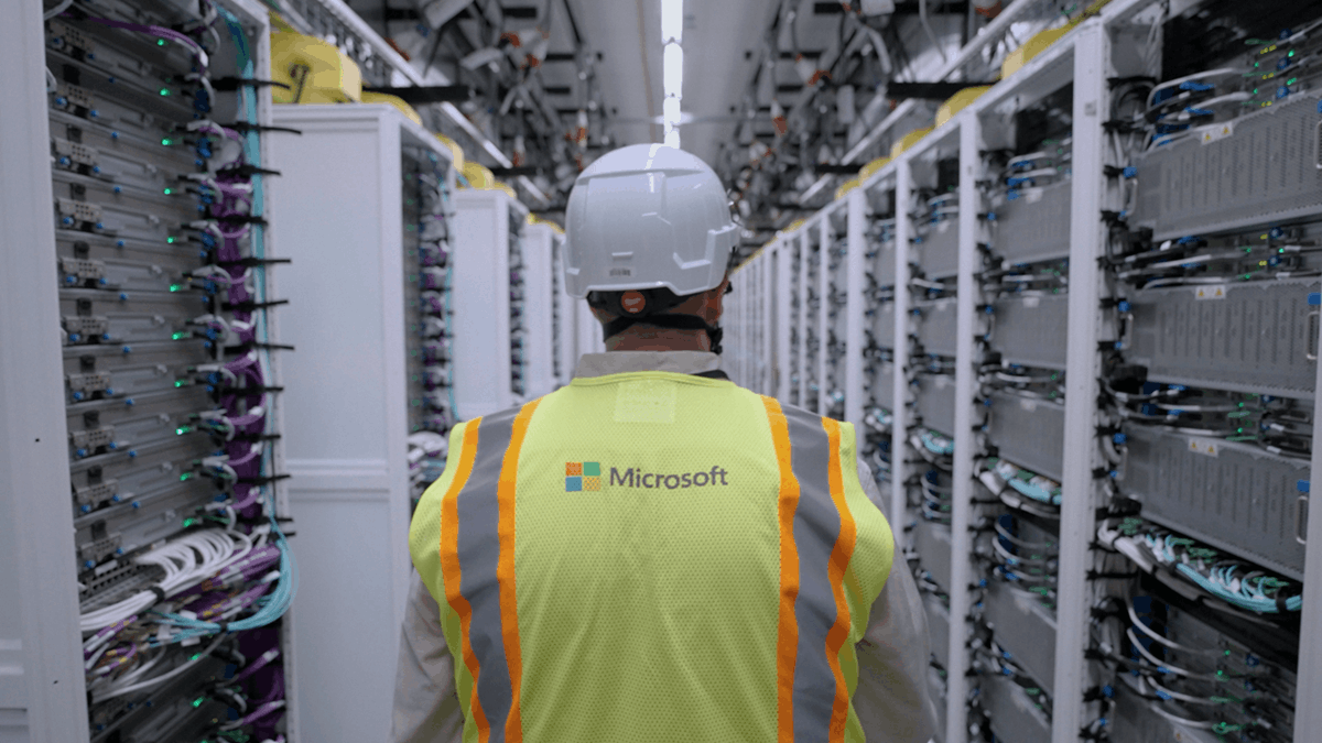 A technician wearing a Microsoft safety vest walks through a data center aisle lined with servers and networking equipment