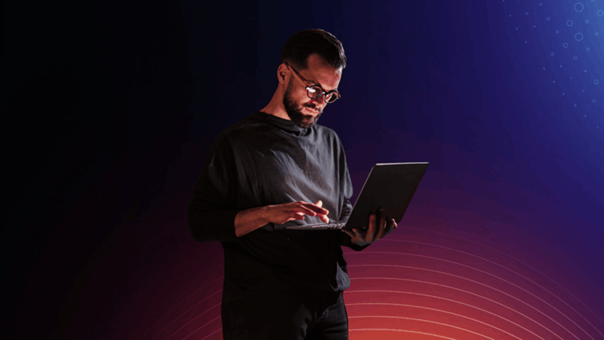 A man using a laptop in a dark setting with blue and red gradient lighting, representing AI and cybersecurity work