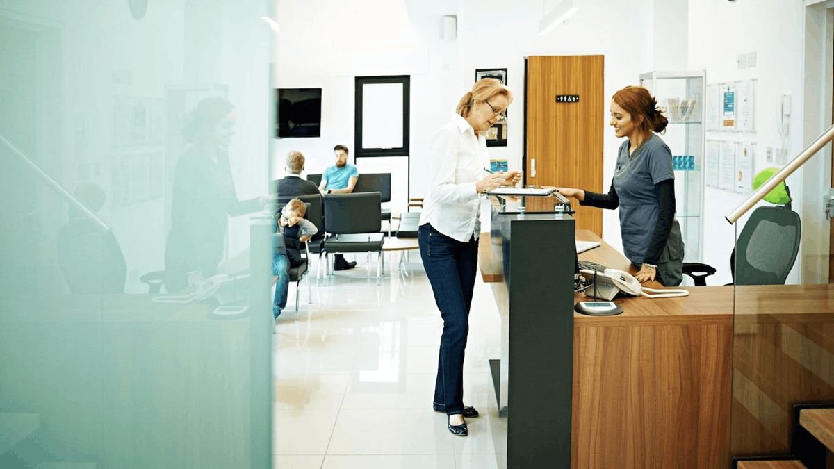 A healthcare receptionist assisting a patient at a clinic front desk while other patients wait in the reception area