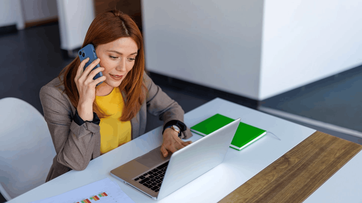 Professional woman speaking on a smartphone while working on a laptop at an office desk